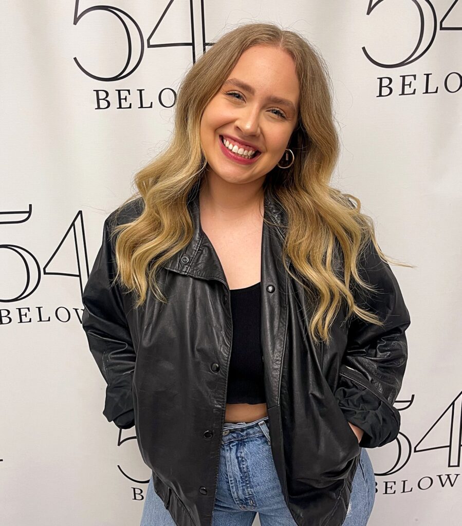 A young woman smiles warmly while posing in front of a step-and-repeat banner for 54 Below, She wears a black leather jacket over a black crop top and light blue jeans