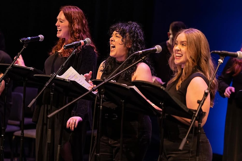 Three women stand side-by-side on stage, each holding a script at a music stand and singing into individual microphones. They wear black attire under stage lighting.