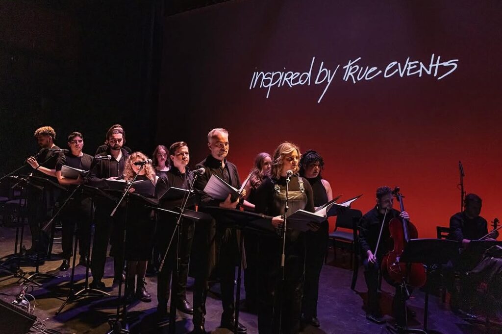 A large ensemble of performers stands in a row on stage, each at a music stand with scripts, singing into microphones. They are dressed in black and lit by stage lighting. Behind them, the words “inspired by true events” are projected onto a red-to-black gradient backdrop.