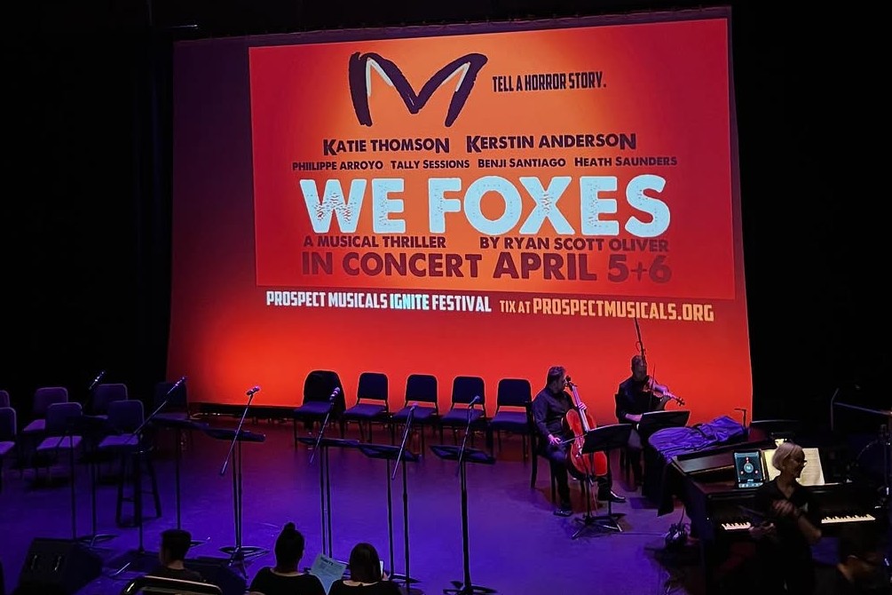 A wide shot of the We Foxes: in Concert stage before or after the performance. The backdrop features the show’s title, dates, and production details in bold red and white text. Music stands and chairs are arranged in front of the projection, with musicians seated in the orchestra pit preparing their instruments.