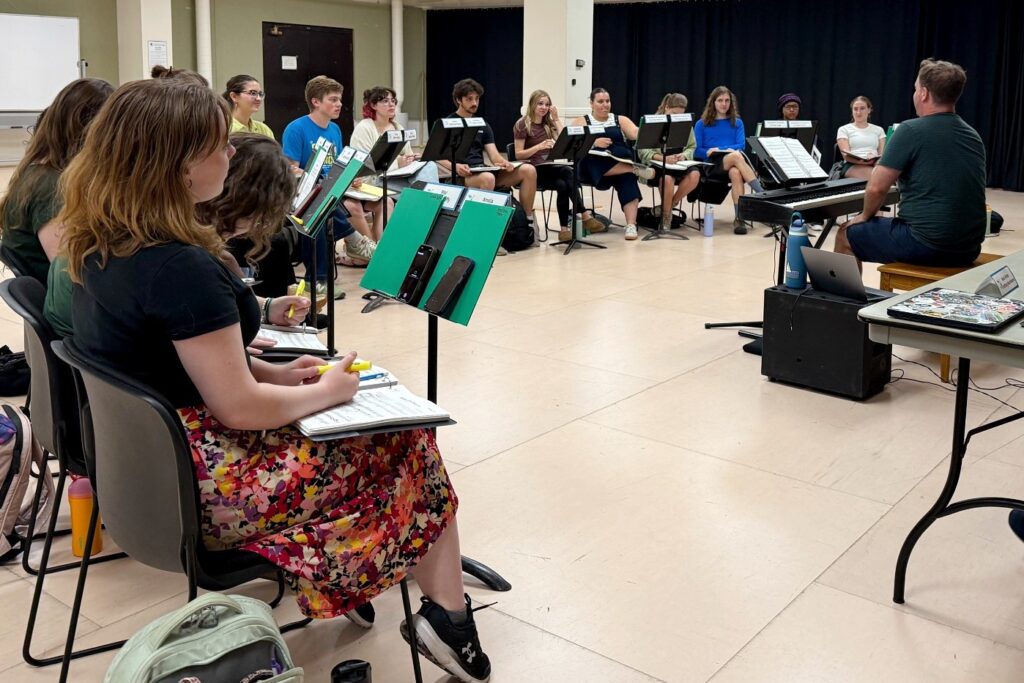 A group of students sit in a semicircle in a rehearsal room, each with a music stand, scripts, and highlighters in hand. The instructor, seated at an electric keyboard with a laptop nearby, faces the group.