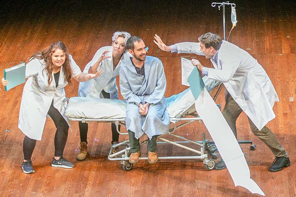 Three actors are dressed in scrubs and surround another actor in a hospital gown sitting on a gurney with an attached IV pole.