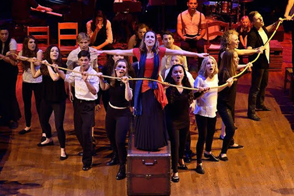 A cast of actors on a stage in a theatre setting. The actor in the center stands on a steamer truck. The other actors around her form a V that is defined by the long rope they all hold.