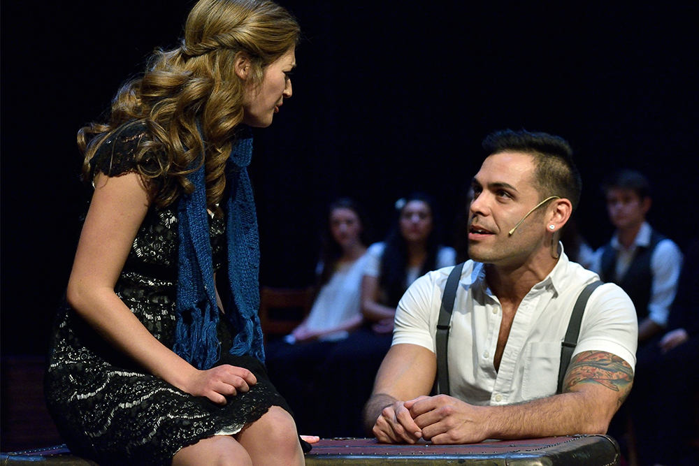 Two actors on stage. The woman on the left sits on a steamer trunk. She wears a sequined dress and a knit scarf and sings to the other actor sitting alongside the trunk. He wears a white collared shirt and suspenders.