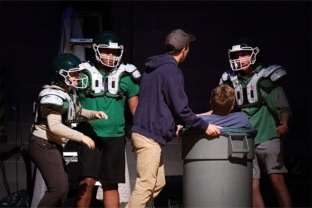 Five actors are on stage with three of the actors dressed in football pads and helmets. One of the actors holds the edge of a trash can that another actor is sitting in.