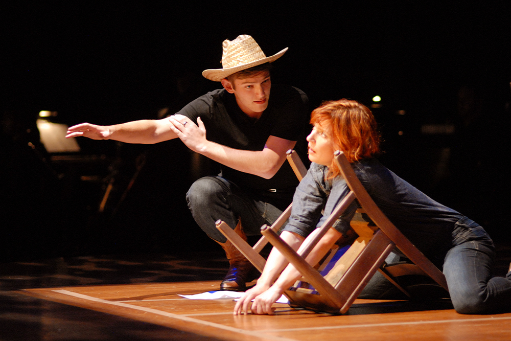 Two actors are on stage in a theatre setting. One actor wears a black shirt, straw cowboy hat, and cowboy boots. He is crouching and gestures out away from the other actor. The other actor is sitting with her arms outstretched through the bottom of an upturned wooden chair. She is wearing a denim shirt and jeans.