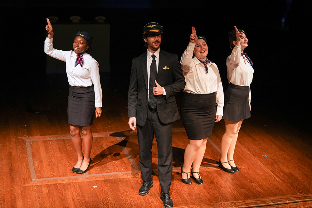 Three actors in flight attendant costumers with dark skirts, white shirts, hats and scarves tied around their necks stand around an actor in a dark suit with a piolet hat and aviation wings pin. The actors are on a wooden stage in a theatre setting.