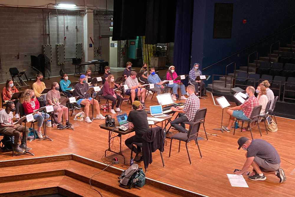 A group of performers sit on stage behind music stands. The are facing writers and directors at a table and in chairs. Everyone is on a stage in a theatre setting.