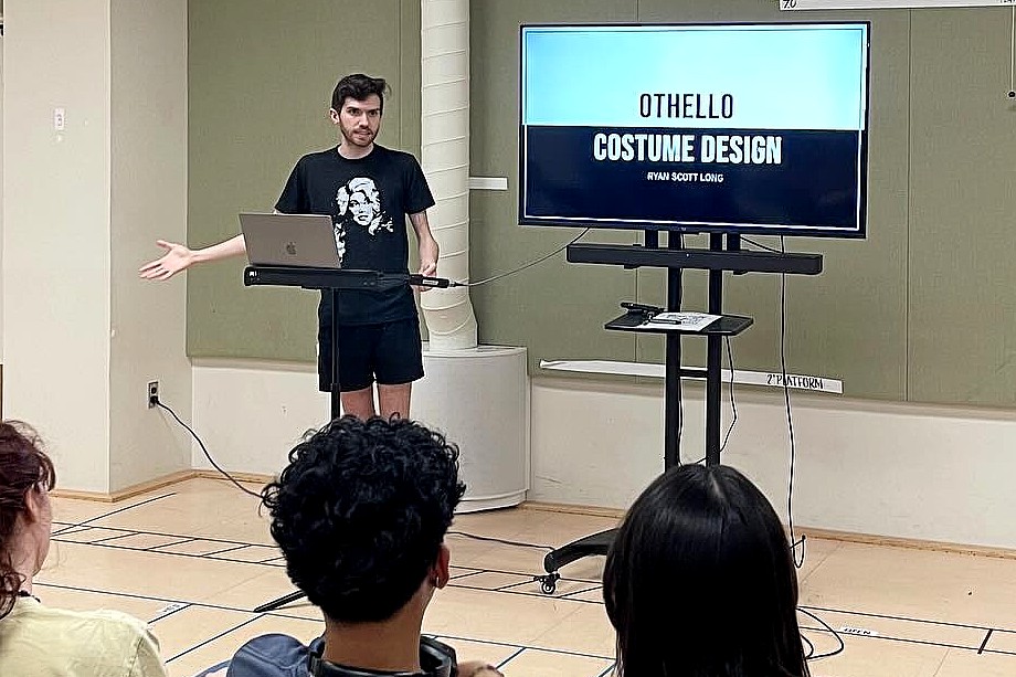A student stands at the front of a classroom presenting a slideshow titled “Othello Costume Design” while classmates seated in rows watch attentively.