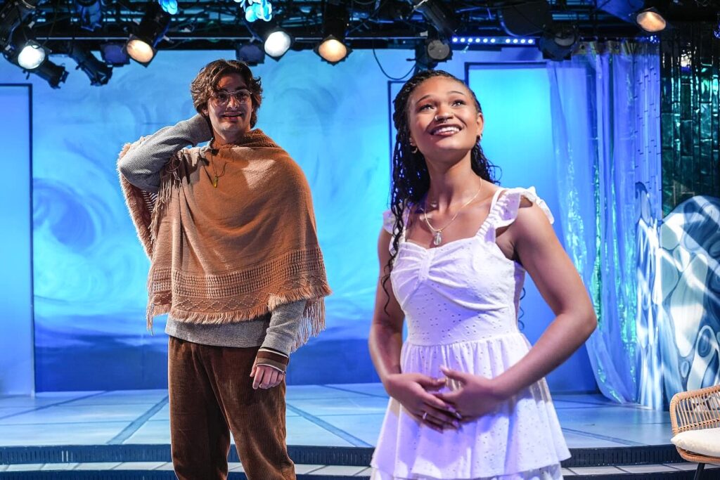 Two actors stand on a brightly lit stage with a blue, wave-patterned backdrop. The actor on the left wears a light brown poncho over a gray sweater and corduroy pants, while the actor on the right, in a white eyelet dress, looks upward.