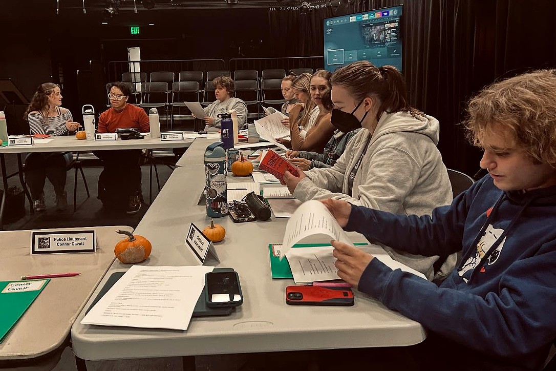 Students sit around a large rectangular table in a black box theater, reading from scripts and notes. Nameplates, small pumpkins, and water bottles are placed in front of them.