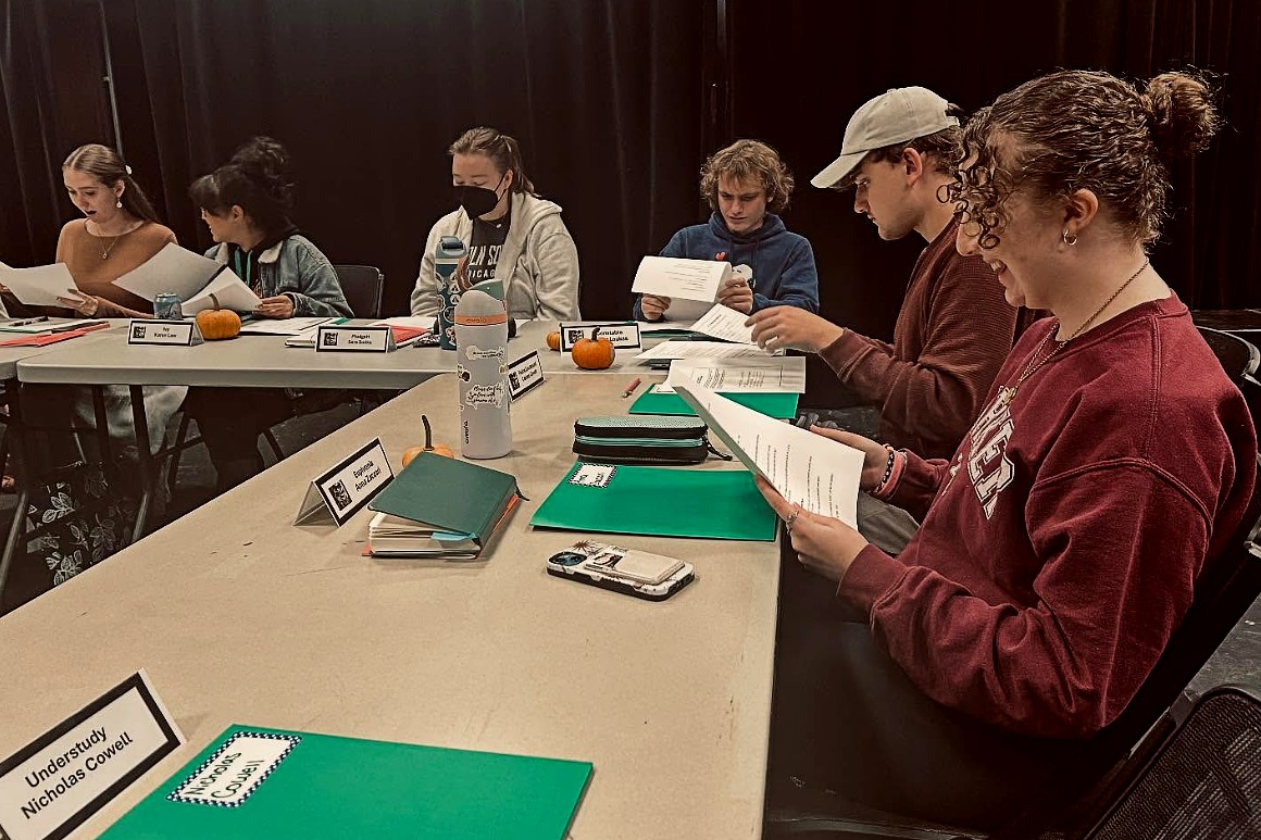 A group of students sit at a long table covered with green folders, papers, and nameplates, reviewing scripts together in a rehearsal or meeting setting.