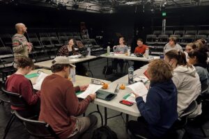 A person stands and speaks to a group of students seated around tables arranged in a square inside a black box theater. The students hold scripts and listen attentively, surrounded by nameplates and small pumpkins.