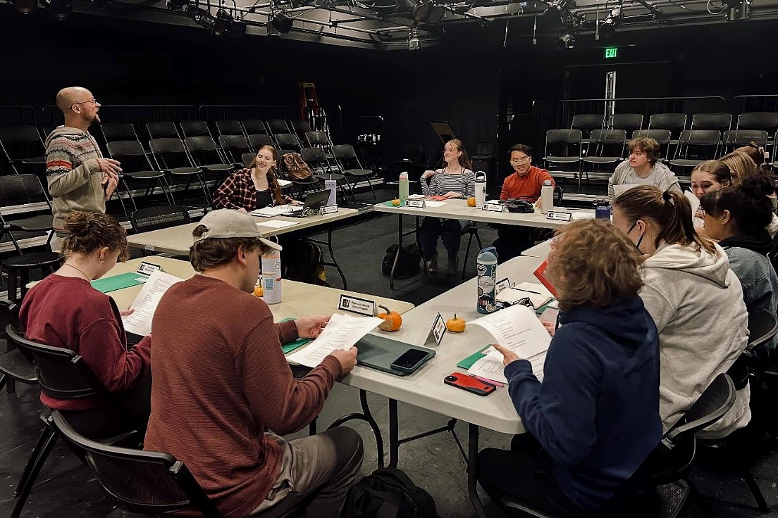 A person stands and speaks to a group of students seated around tables arranged in a square inside a black box theater. The students hold scripts and listen attentively, surrounded by nameplates and small pumpkins.