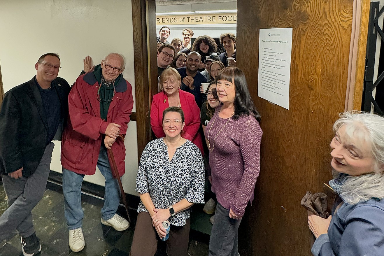 A mixed group of people gathers in a hallway outside the door to a room labeled “Friends of Theatre Food Pantry.” Some people stand in the hallway while others lean in through the open doorway from inside the pantry. 