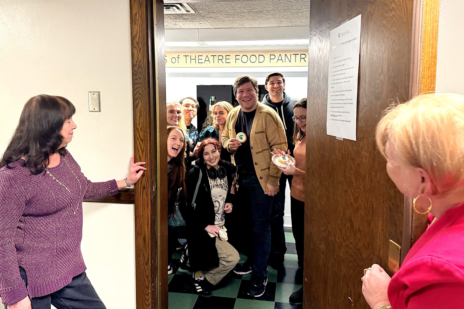 Several people crowd together inside a small room marked “Friends of Theatre Food Pantry,” smiling toward the camera through a doorway. Two adults stand on either side of the open door, looking in. 