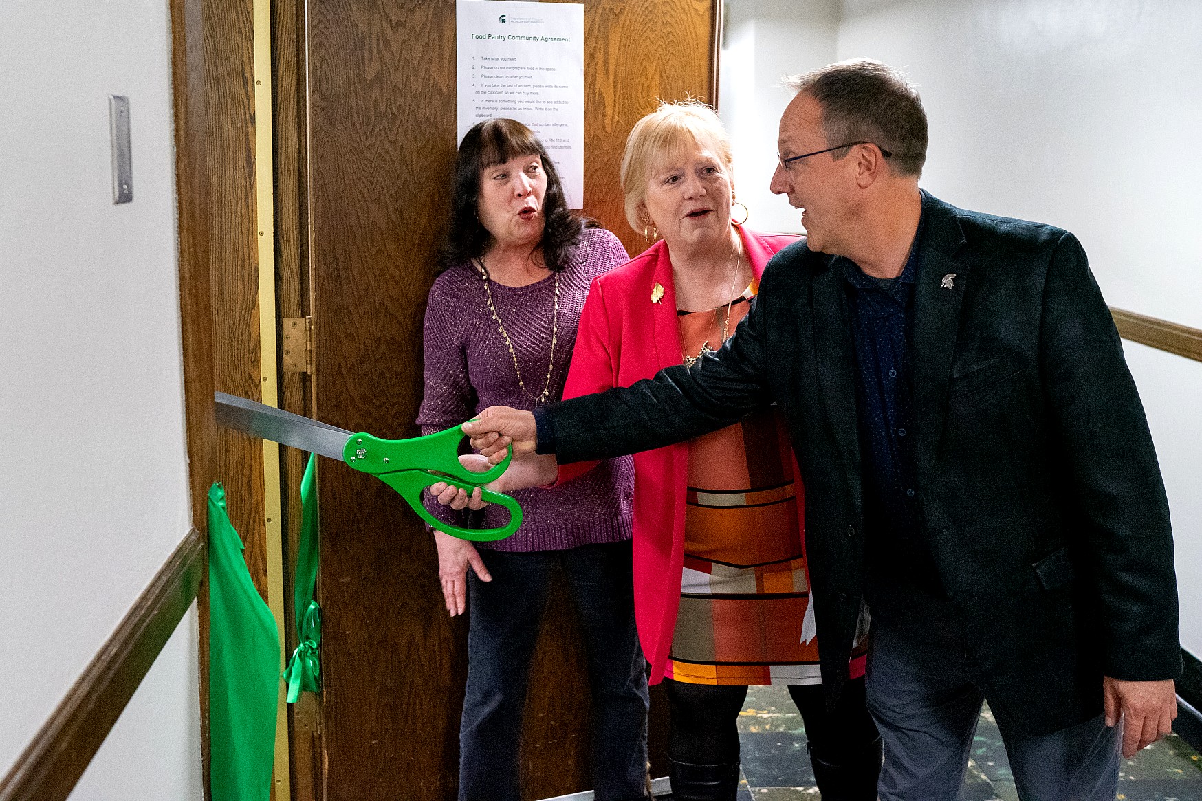 The same three individuals stand in the hallway at the ribbon-cutting site. One person holds the oversized green scissors extended toward the ribbon, while the two others stand close behind.