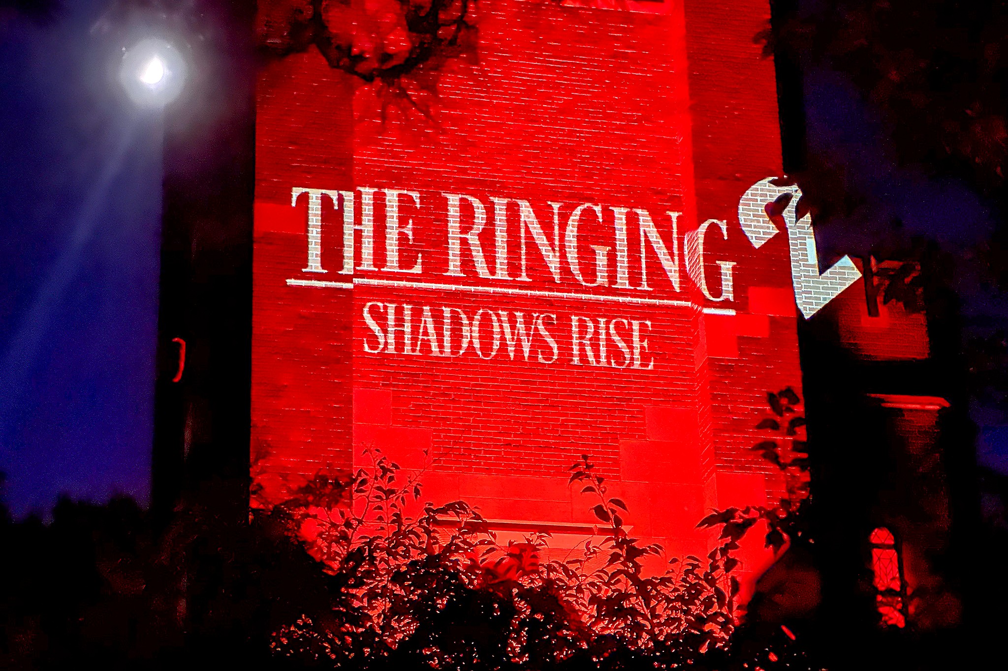 A close-up view of Beaumont Tower glowing red with the projected words “The Ringing: Shadows Rise,” partially framed by silhouetted branches.