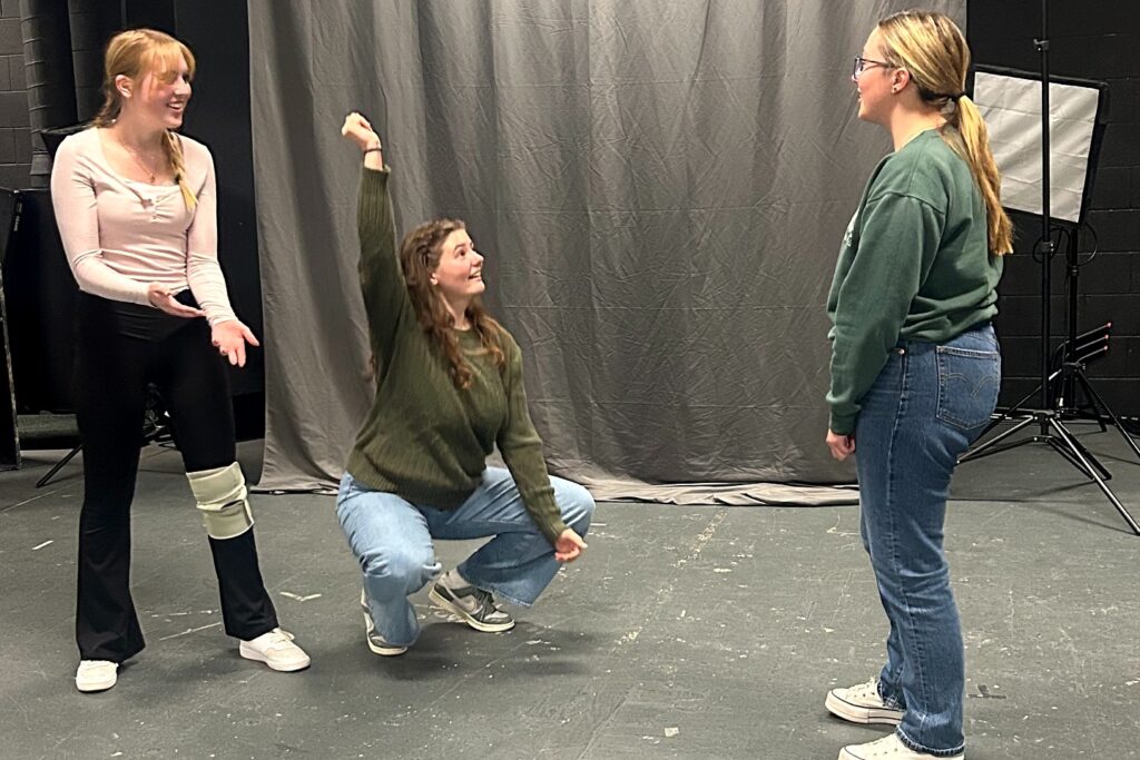 Three students participate in an improv activity in a studio. One stands smiling with hands open, another crouches with an arm raised, and a third faces them while listening.