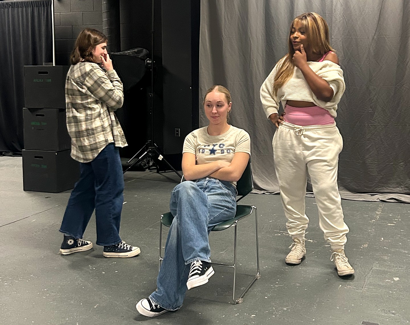 A seated student with folded arms looks ahead as two standing students observe her, each with a hand on their chin.
