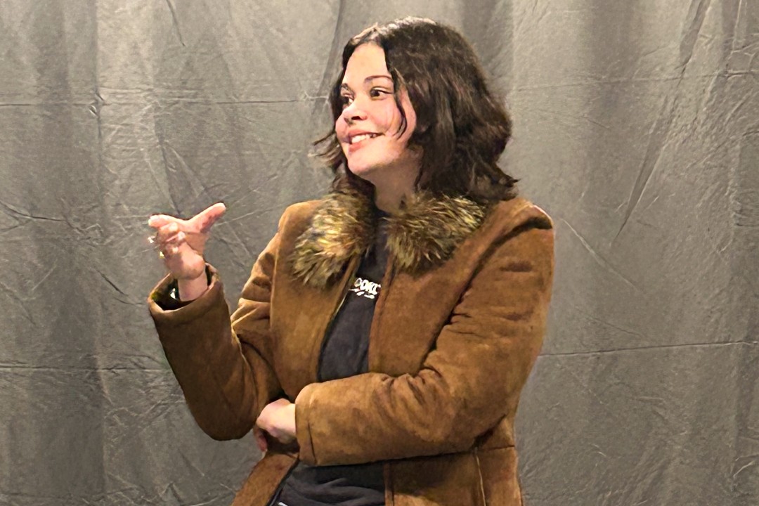 A student in a brown coat with a fur-trimmed collar gestures with one hand while smiling during an improv scene. 