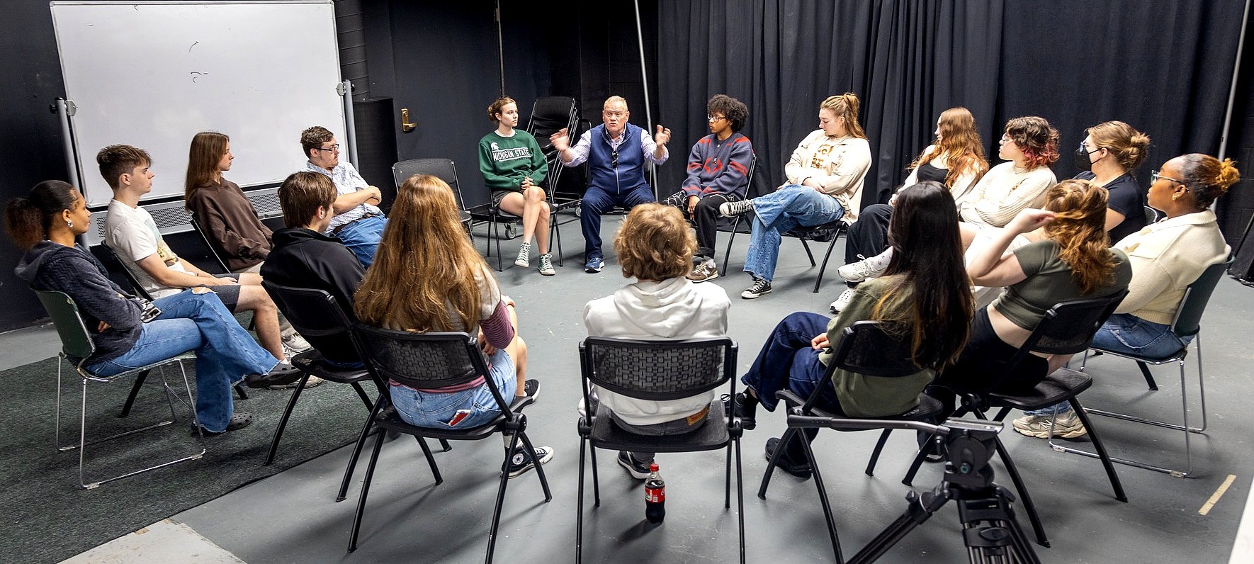 Larry Joe Campbell leads a discussion with a group of approximately 15 MSU Department of Theatre students seated in a large circle. The session takes place in a theater rehearsal space with a whiteboard and black curtains in the background.