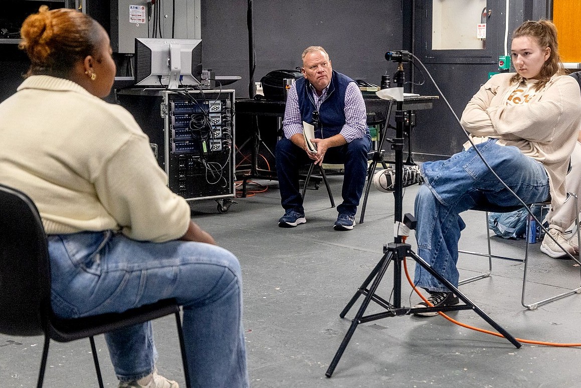 Larry Joe Campbell sits in the background, observing a student during a scene rehearsal. The foreground shows a student sitting in a chair facing away from the camera.