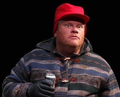 Larry Joe Campbell performing in the 2010 Central Michigan University alumni production of Almost Maine. He is wearing a red hunting cap, a patterned heavy coat, and black gloves while holding a beverage can.