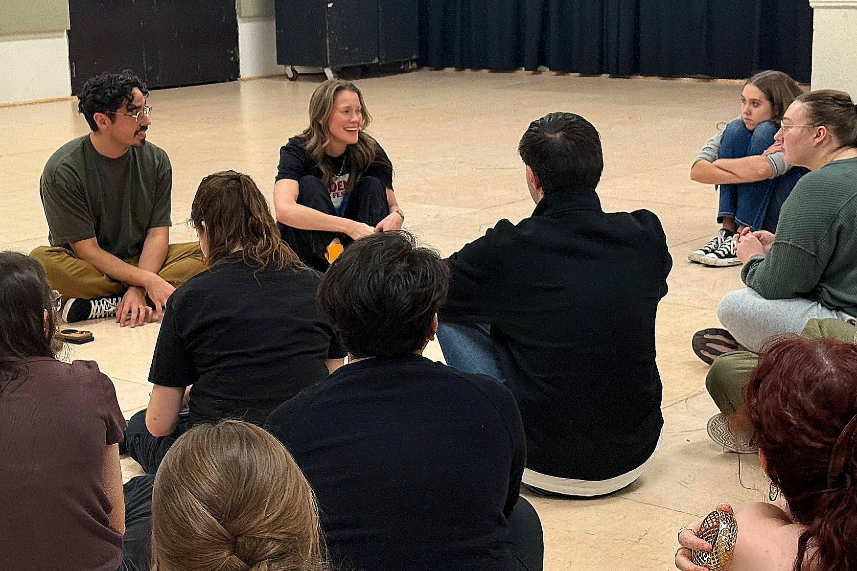 A group of people in different styles of dress sit on the floor of a brightly lit rehearsal studio.