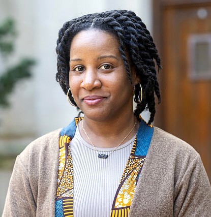 A professional headshot of Alexandria Davis, Assistant Professor of Dance at Michigan State University. Her hair is styled in twists, and she wears gold hoop earrings, a thin gold necklace with a dark pendant, and a tan cardigan over a ribbed grey top with a blue and yellow patterned trim. She is looking toward the camera with a neutral, poised expression.