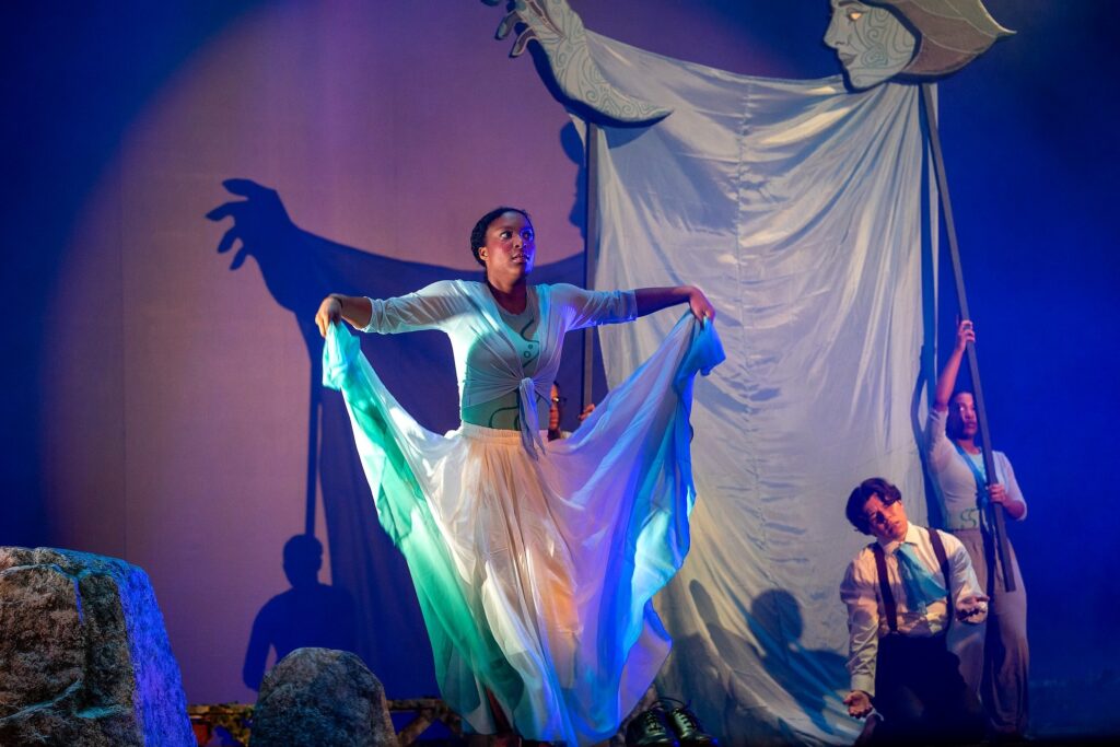A dancer stands center stage holding flowing blue-green fabric that cascades like water, illuminated by cool lighting. Behind her, a large sculptural figure with outstretched arms casts a dramatic shadow, while another performer kneels nearby.