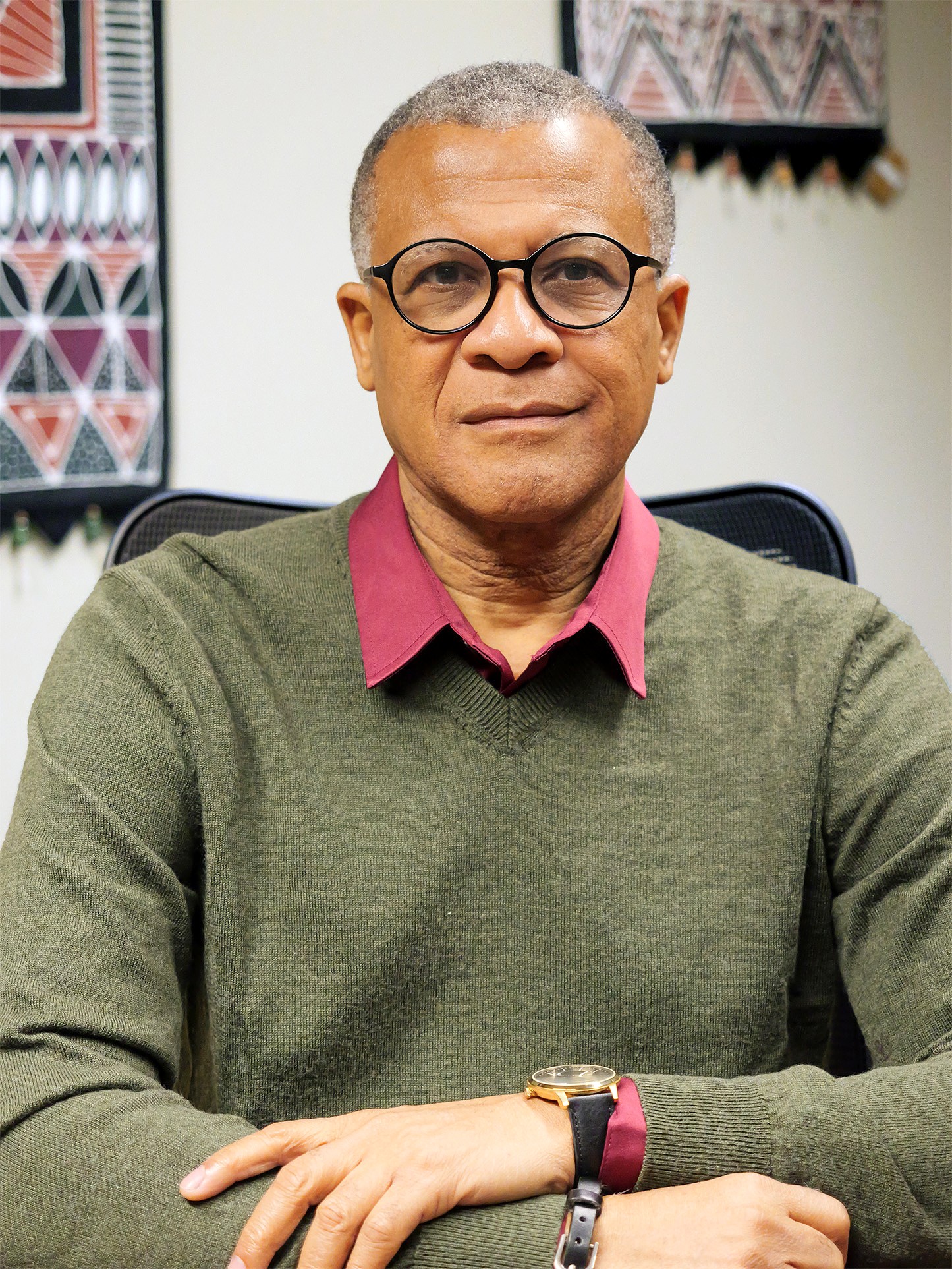A professional headshot of Philip Effiong, Professor of Theatre Studies and Integrative Humanities. He is wearing round black-rimmed glasses, an olive green V-neck sweater over a maroon collared shirt, and a watch with a black strap.