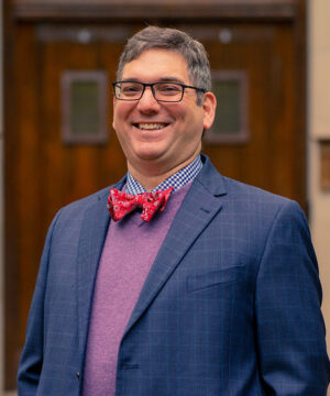 Stephen DiBenedetto Stands Before Beaumont Tower Wearing Glasses and Smiling in a Blue Checked Jacket Purple Sweater and Red Bowtie
