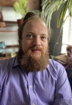 Man with blond beard and blue eyes, wearing a purple and white gingham button down, collared shirt sits with plants and shelving in the background