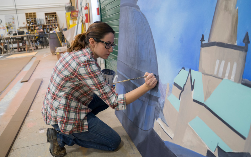 woman wearing glasses and a flannel kneeling down painting a backdrop