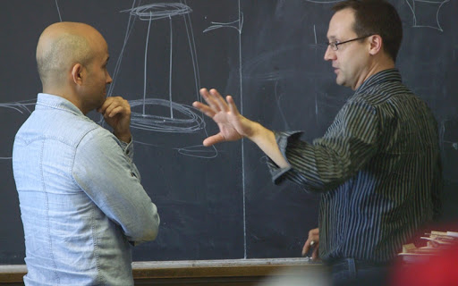 two men standing in front of a chalkboard one explaining what the drawings mean