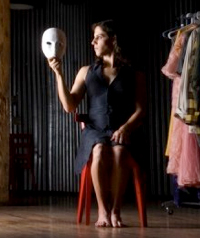 A woman in a dark dress holds up a theatrical mask.