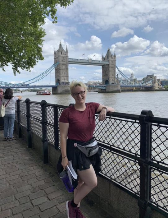 Woman with blonde hair wearing a red t-shirt and black shorts is leaning against a black fence in front of the London Bridge