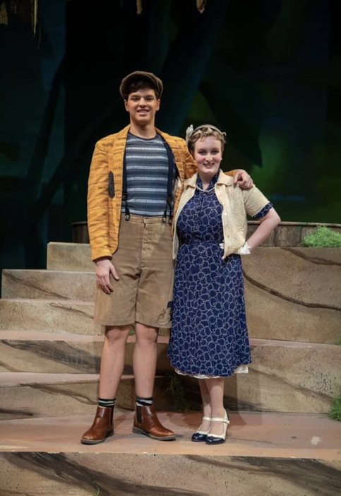 A man (left) and a woman (right) standing on a stage. The man is wearing a yellow jacket, striped shirt, cargo shorts, and a hat. The woman is wearing a blue ankle-length dress and a tan cardigan.