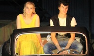 actor in a yellow dress sitting next to an actor in a tee, in a car