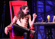 an actor in only pants is sitting in a big red chair, looking to an end table on his right