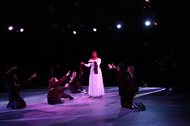 actor in a white down standing in the center, actors dressed in black at kneeling for her