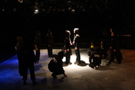 two actors dressed in black standing in the center, other actors are dressed in black and crouched, all actors are obscured