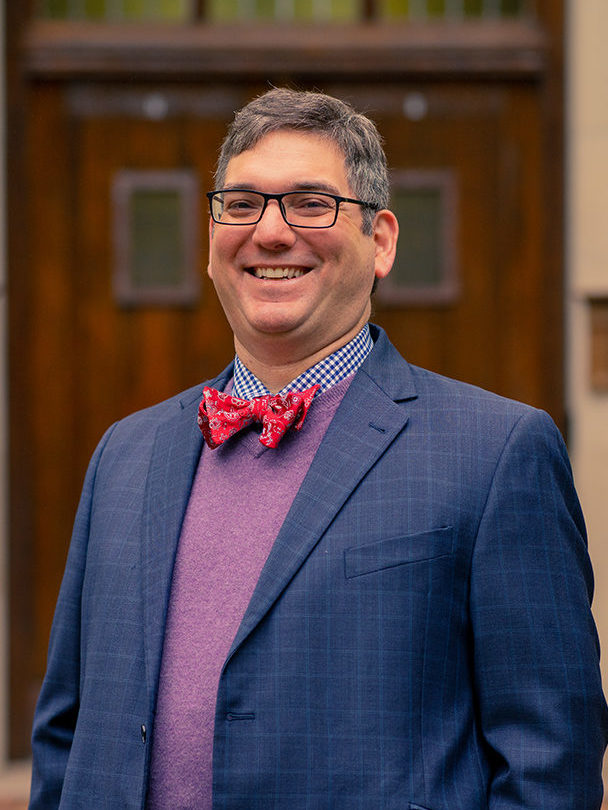 Stephen DiBenedetto Stands Before Beaumont Tower Wearing Glasses and Smiling in a Blue Checked Jacket Purple Sweater and Red Bowtie