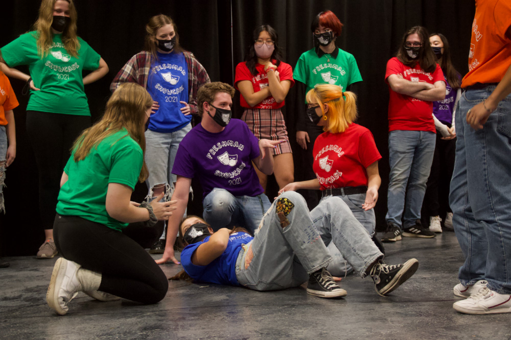 Students rehearse for the improv comedy show. One student lies on the ground, surrounded by three kneeling classmates. Other students watch in the background