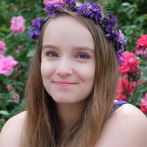 Headshot of a person wearing a purple flower crown. In the background there are pink flowers. 