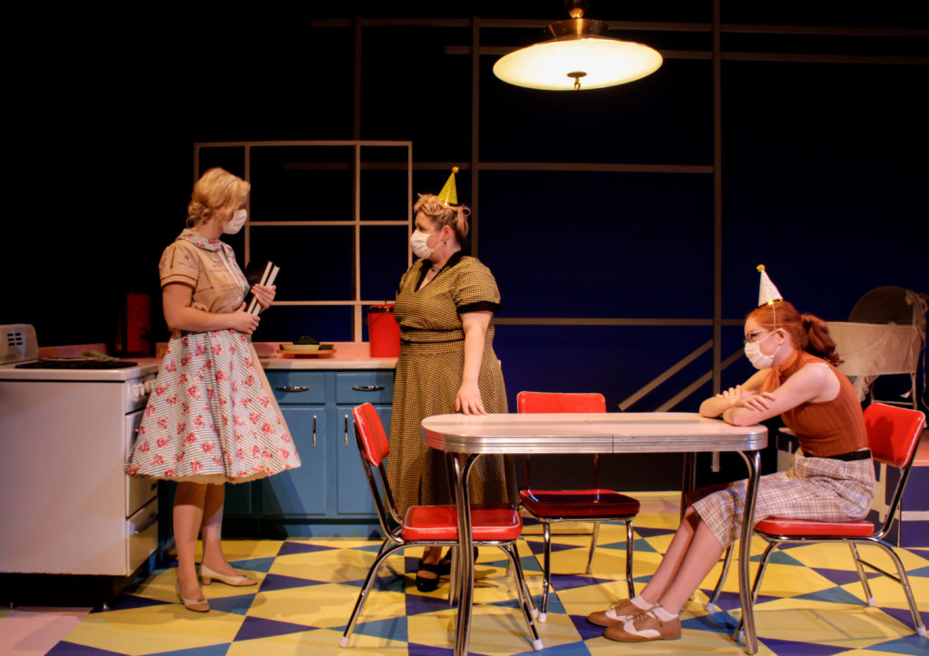 Three actors wearing dresses and masks stand in a kitchen set on the stage.  