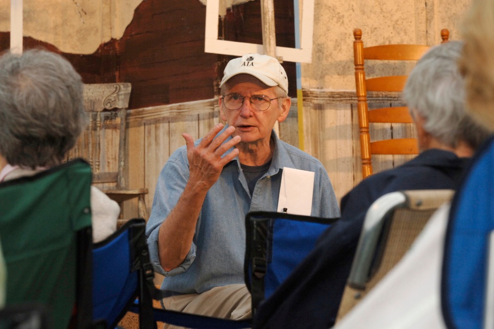 an older ma wearing glasses, a white hat and blue button down shirt talking to a group of people