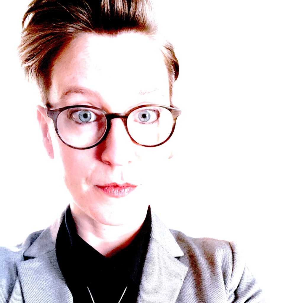 A picture of a woman with short red hair and glasses looking at the camera. She wears a grey blazer and black turtleneck.