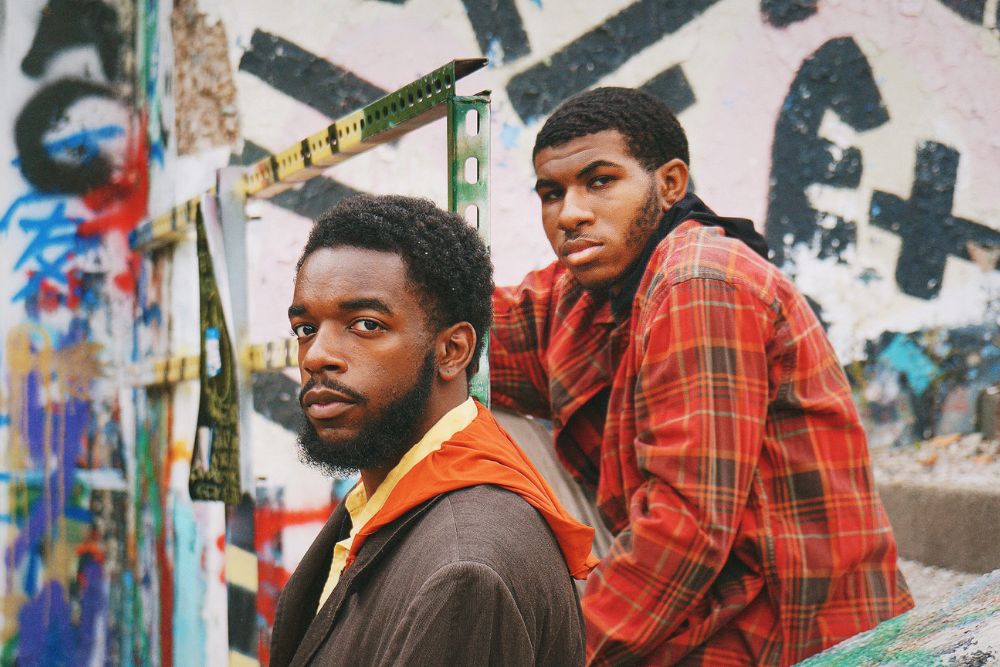 Two men outdoors in a graffiti covered area looking into the camera.